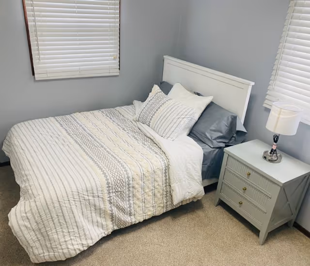 A neatly made bed with a white and gray patterned comforter and multiple pillows in a small bedroom. The bed has a white headboard and is positioned between two windows with white blinds. Next to the bed is a light gray nightstand with three drawers and a small lamp on top. The walls are painted light gray and the floor is carpeted.