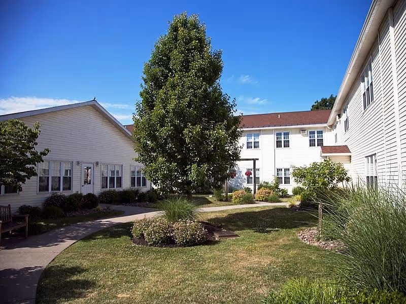 Sunlit courtyard featuring a central leafy tree, grassy beds, and two-story white buildings with a curved walkway.