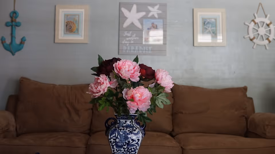 Blue-and-white vase of pink flowers on a table in front of a brown couch with nautical wall decor.