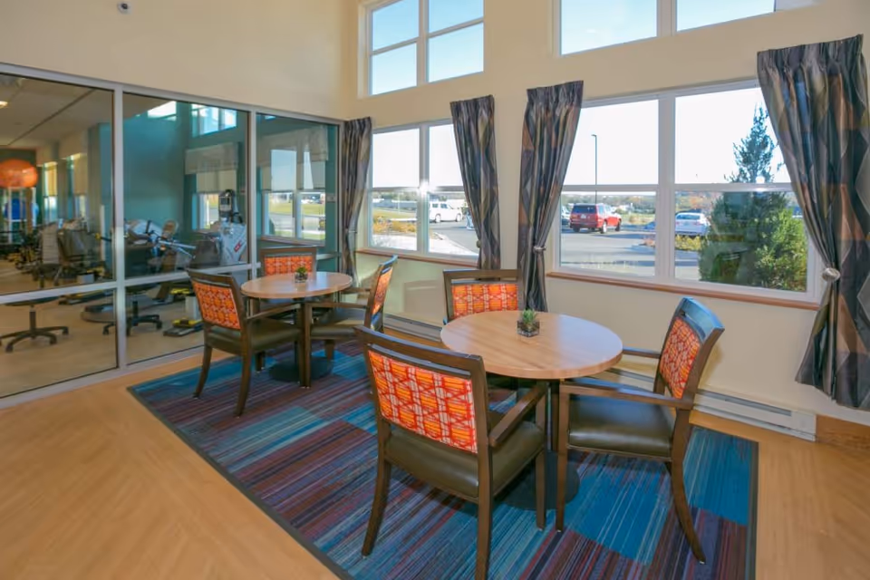 Bright communal seating area with round tables and patterned chairs on a striped rug beside large windows and a glass wall revealing an exercise room.