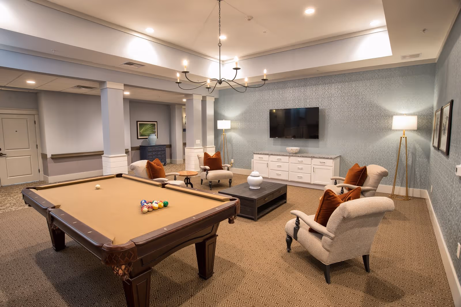 A cozy lounge area in a senior living facility featuring a pool table with balls arranged for a game, four cushioned armchairs with orange pillows arranged around a dark wooden coffee table, a white cabinet with a flat-screen TV mounted above it, two floor lamps, and soft patterned wallpaper on the walls.