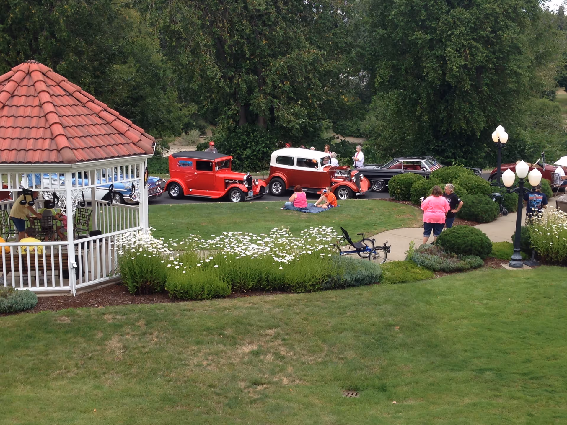 A grassy outdoor area with a white gazebo, people seated on the lawn, and several vintage red cars parked along a driveway.