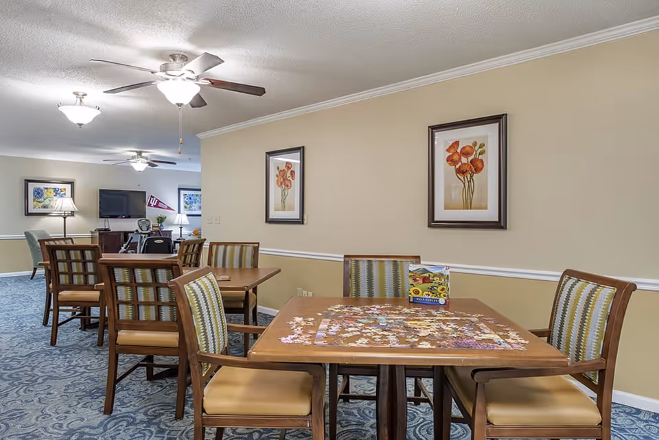 A well-lit common area with several wooden tables and chairs arranged on a blue patterned carpet. One table in the foreground has a partially completed jigsaw puzzle and a puzzle box on it. The walls are beige with white trim and decorated with framed floral artwork. Ceiling fans with lights hang from the white ceiling. In the background, there is a TV mounted on the wall and additional seating.