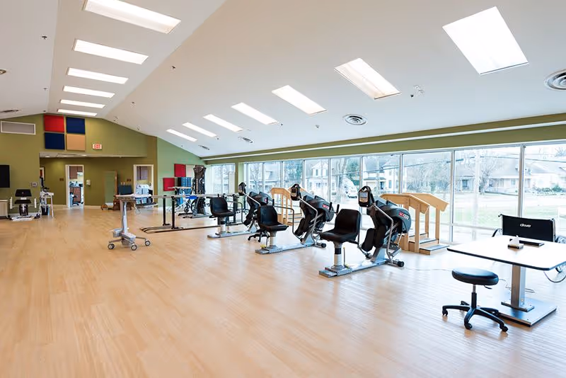 A spacious rehabilitation room with exercise equipment including stationary bikes and parallel bars. The room has large windows letting in natural light, light wood flooring, and a high ceiling with multiple skylights. There are tables and chairs along the right side near the windows.