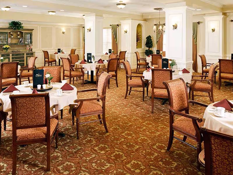 A spacious dining room with multiple round tables covered with white tablecloths, each set with cups, napkins, and menus. The room features patterned carpet, upholstered wooden chairs, cream-colored walls with decorative molding, and soft lighting from ceiling fixtures and wall sconces.