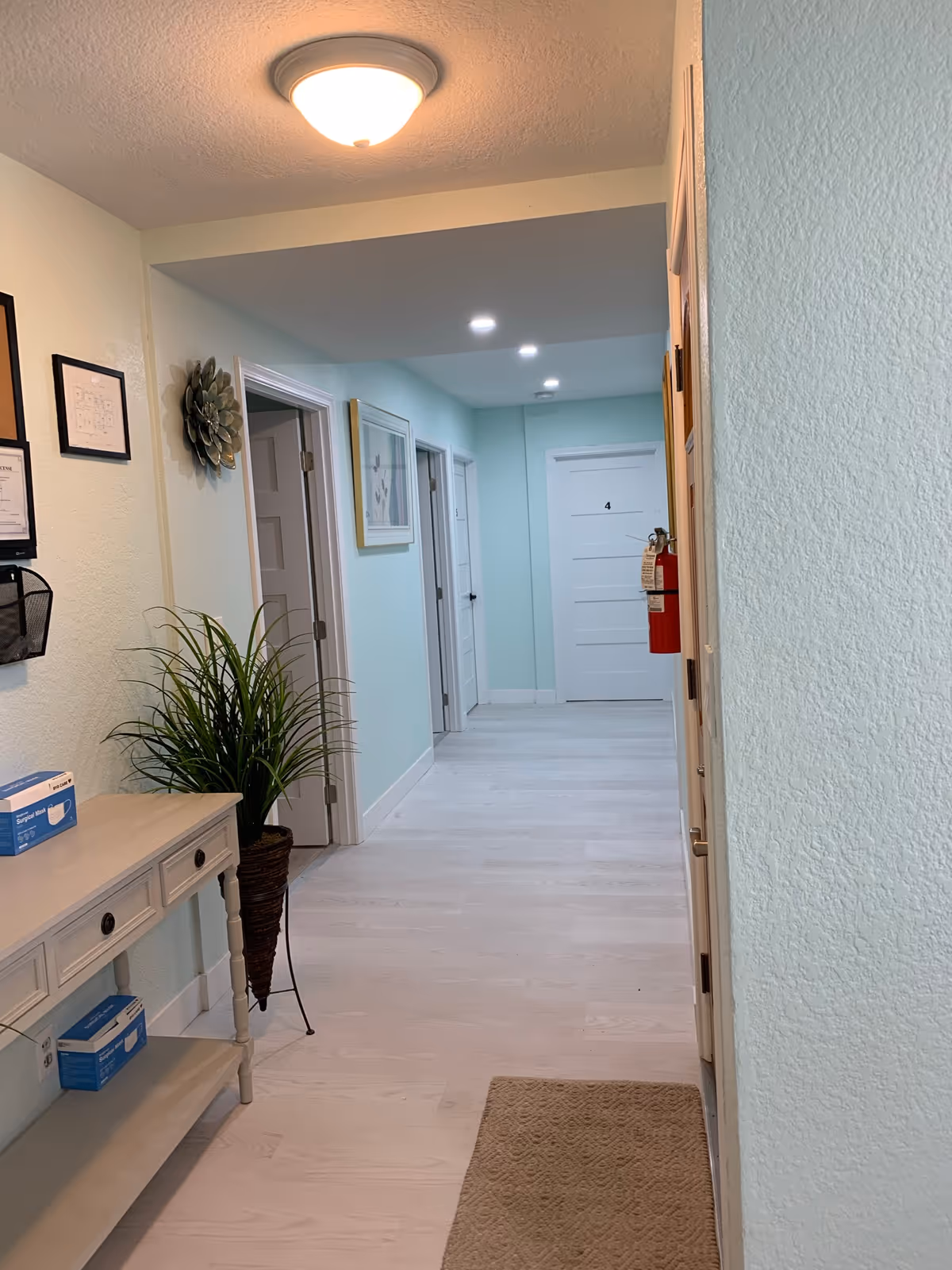 A clean, well-lit hallway in a senior living facility with light blue walls and white doors. The hallway features a beige rug, a small table with drawers holding boxes of disposable gloves, a potted plant, framed pictures on the walls, and a fire extinguisher mounted near the door labeled number 4.