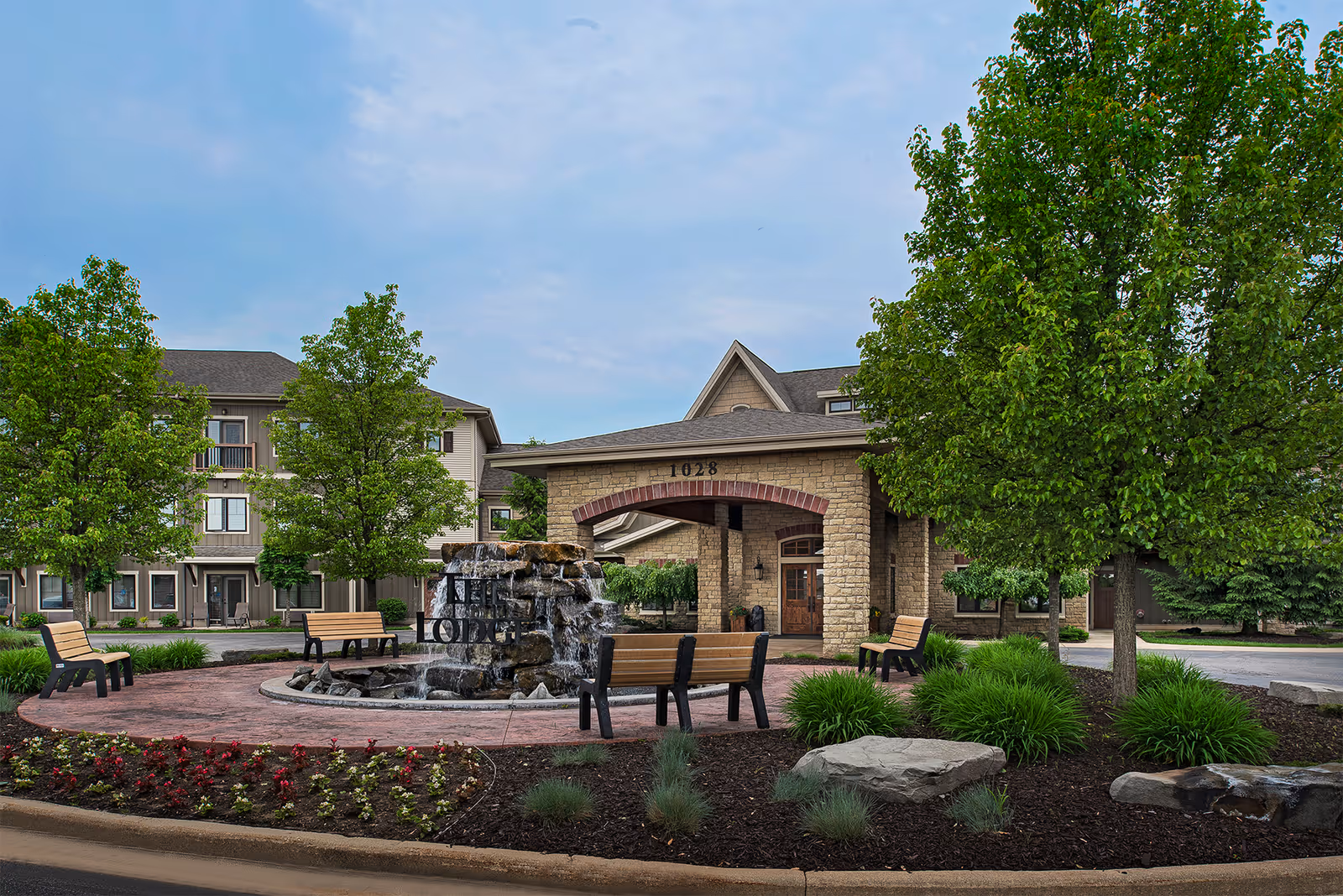 Exterior view of American House Jenison senior living facility showing a stone entrance with an arched roof, surrounded by green trees, benches, a circular water fountain, and landscaped flower beds under a partly cloudy sky.