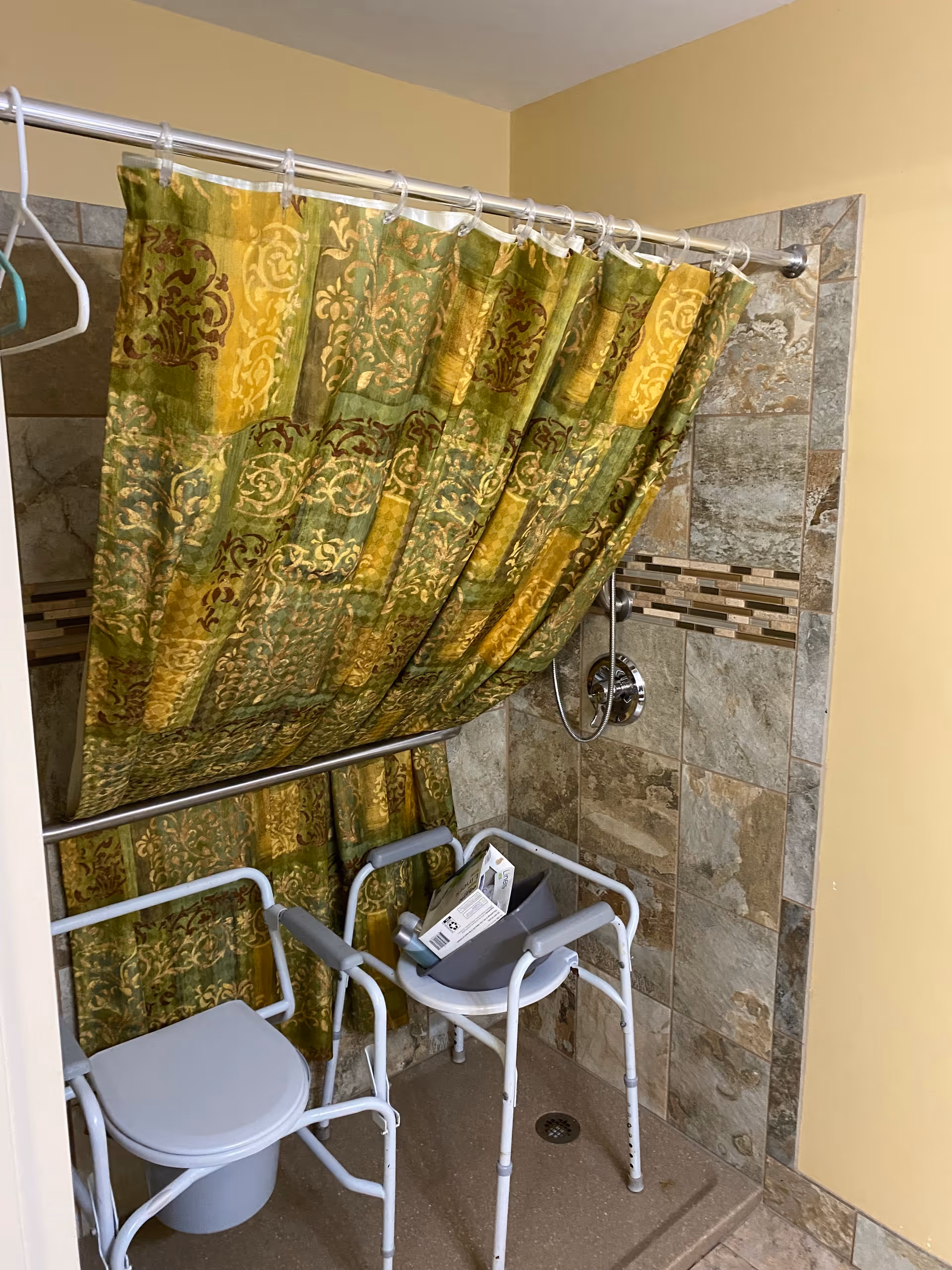 A bathroom shower area with beige and brown tiled walls and a green and yellow patterned shower curtain. Inside the shower are two white metal shower chairs, one with a gray plastic seat and a box placed on it. There is a silver showerhead and a grab bar attached to the wall.