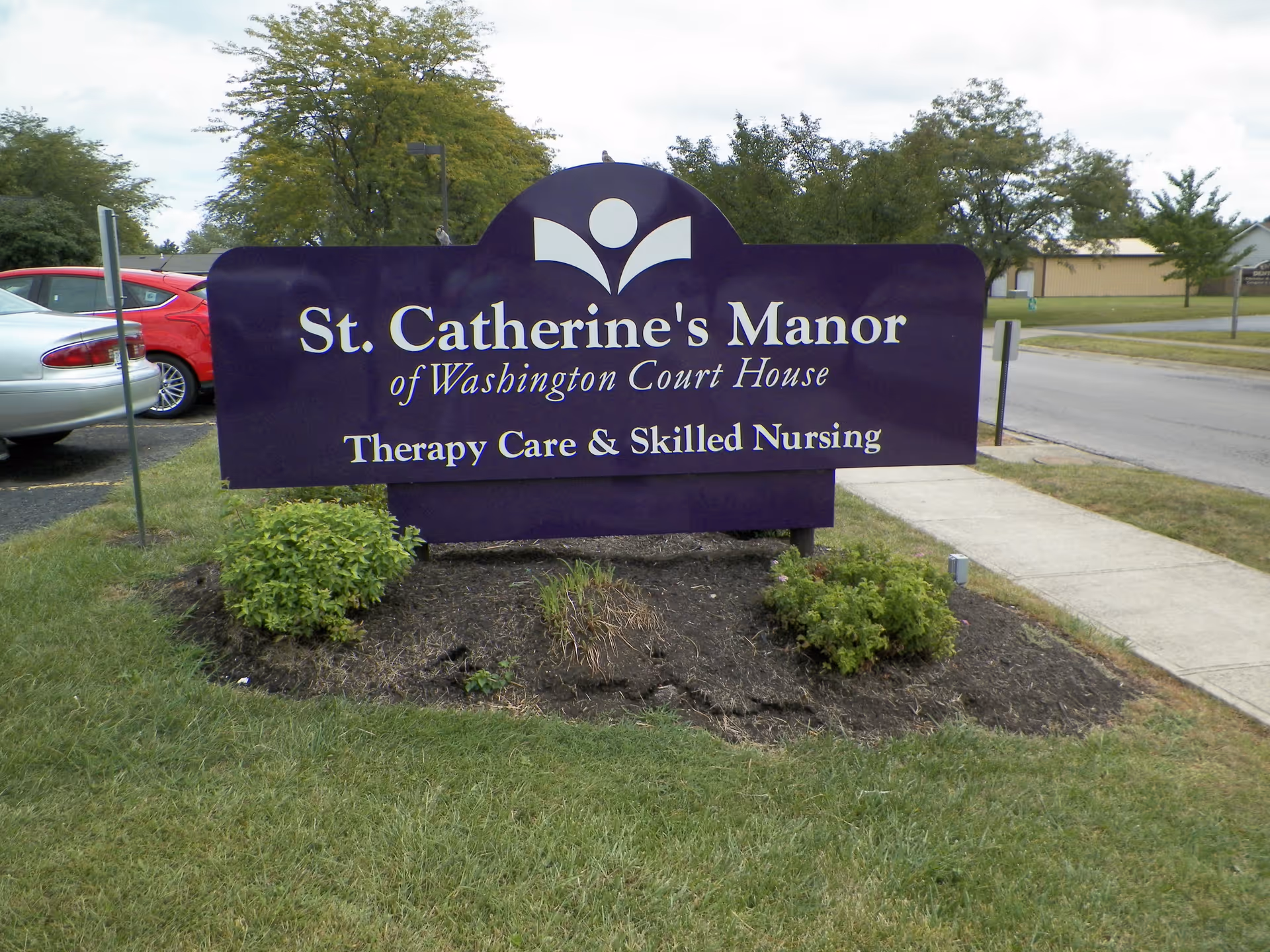 Purple outdoor sign reading 'St. Catherine's Manor of Washington Court House Therapy Care & Skilled Nursing' in a landscaped area by a sidewalk.