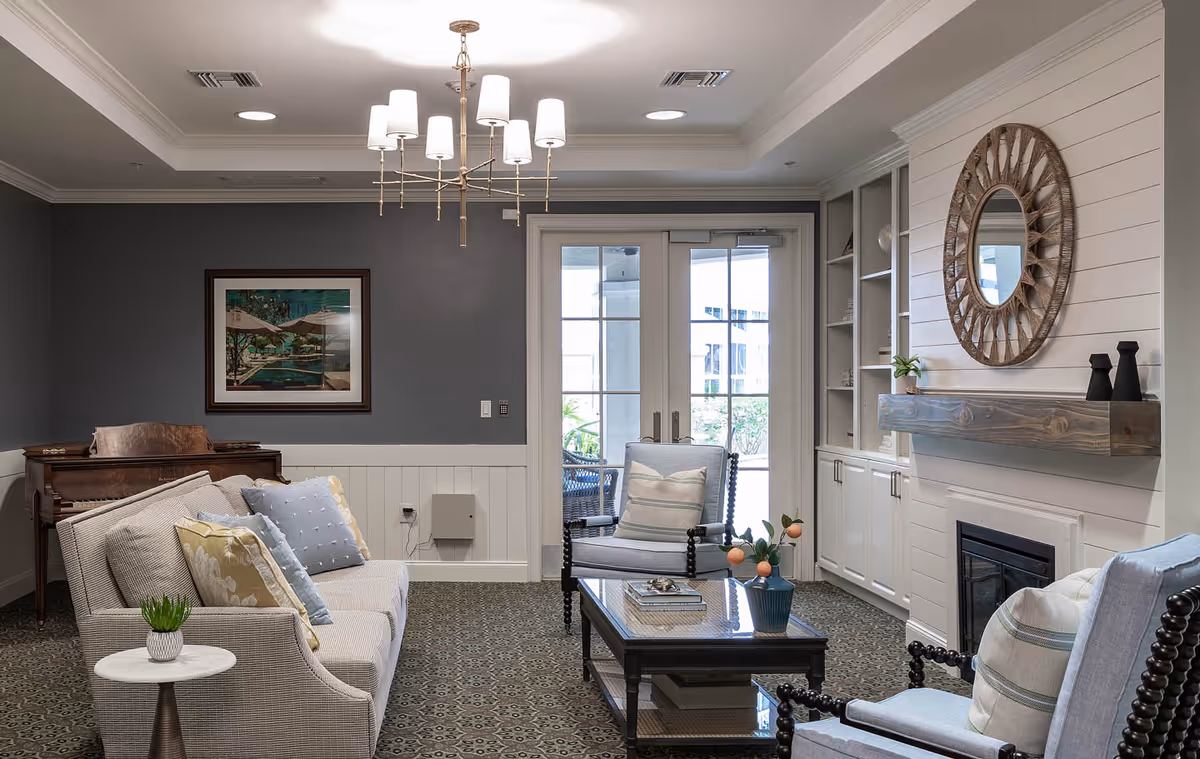 A cozy living room with a light gray sofa adorned with multiple pillows, two cushioned armchairs, a wooden coffee table with decorative items, a fireplace with a wooden mantel and a round mirror above it, built-in white cabinets, a chandelier hanging from the ceiling, and French doors leading outside. There is also a framed picture on the dark gray wall and a piano in the corner.