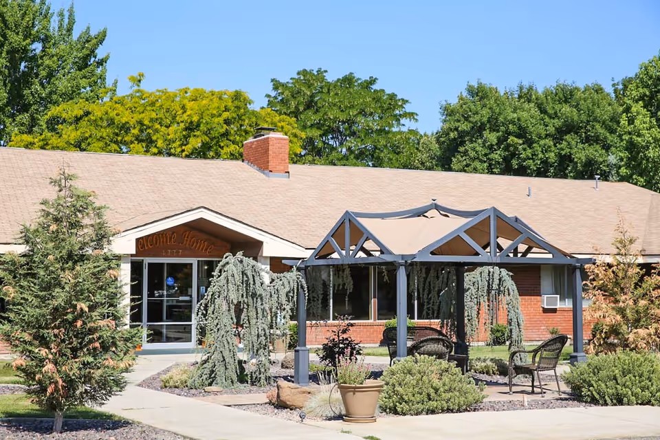 Exterior view of a single-story brick building with a beige roof, surrounded by green trees and shrubs. In front of the building, there is a covered outdoor seating area with a metal frame and a beige canopy, along with several chairs and a potted plant. The entrance has glass doors with a sign above that reads 'Welcome Home'.