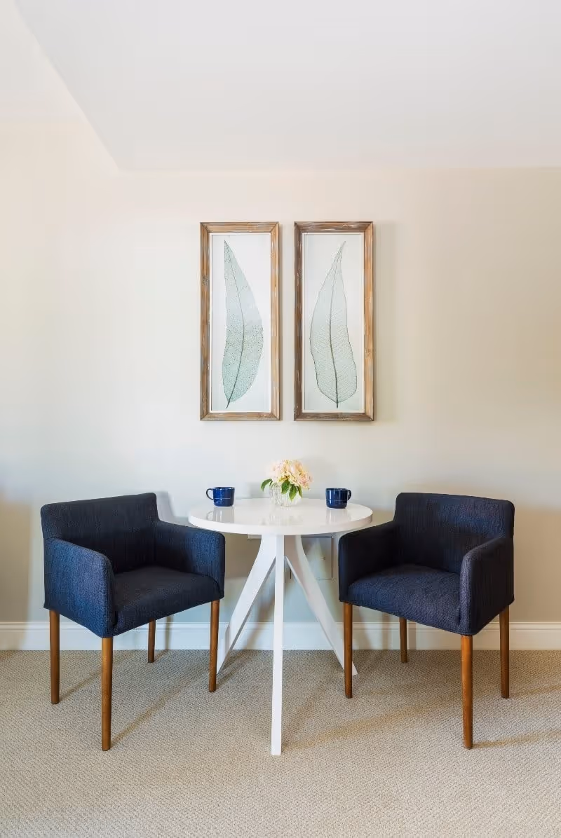 A small round white table with a vase of flowers and two blue mugs on it, flanked by two dark blue upholstered chairs with wooden legs, set against a beige wall with two framed leaf prints hanging above.
