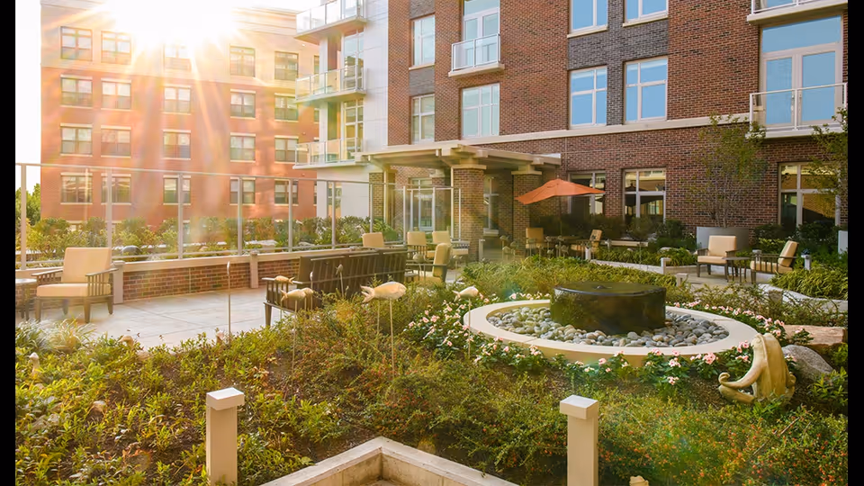 Sunlit outdoor courtyard with a central circular fountain, patio seating, landscaping, and a brick building facade.