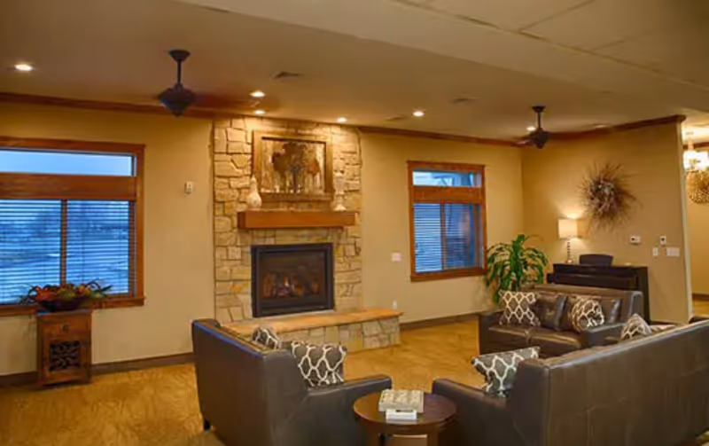 A cozy living room area with a stone fireplace in the center, flanked by two windows with wooden blinds. The room features three dark leather sofas with patterned cushions arranged around a small round table. There are ceiling fans, a potted plant, a lamp on a side table, and warm lighting creating a comfortable atmosphere.