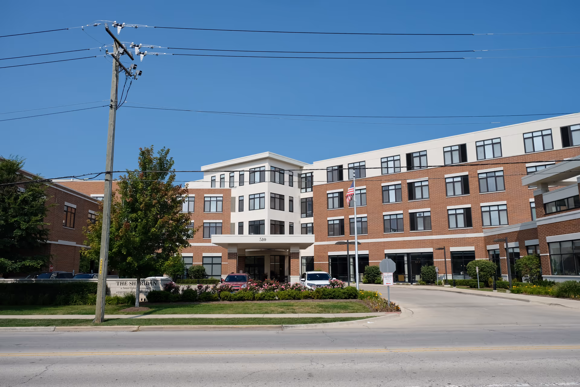 Exterior front view of The Sheridan at Park Ridge, a multi-story senior living facility with a brick and white facade, large windows, an American flag on a flagpole, and a driveway entrance with cars parked near the entrance under a clear blue sky.
