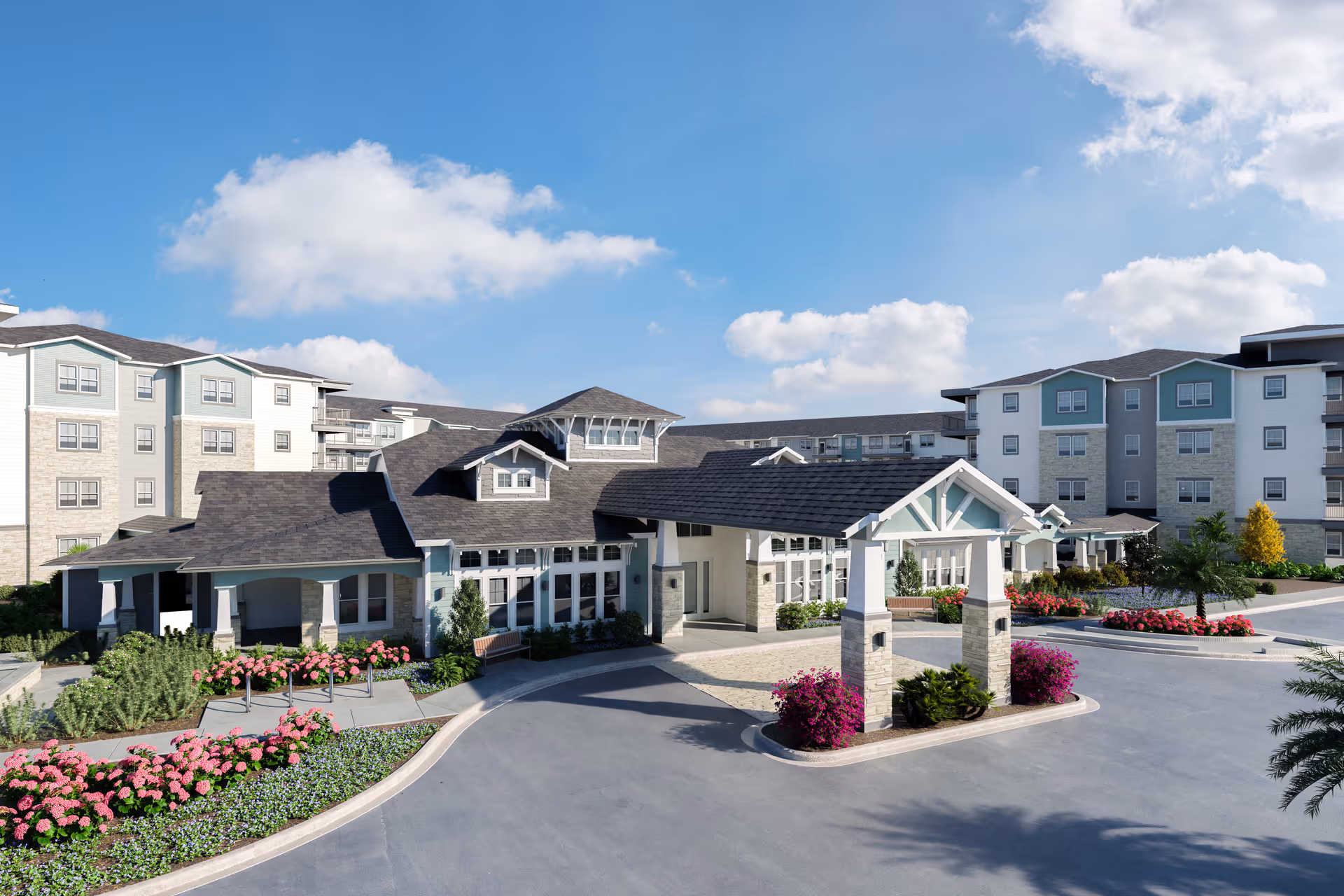 Exterior view of Amberlin South Naples senior living facility showing a large covered entrance with stone pillars, surrounded by landscaped flower beds and bushes. The building has multiple stories with light-colored walls and dark roofs under a blue sky with scattered clouds.