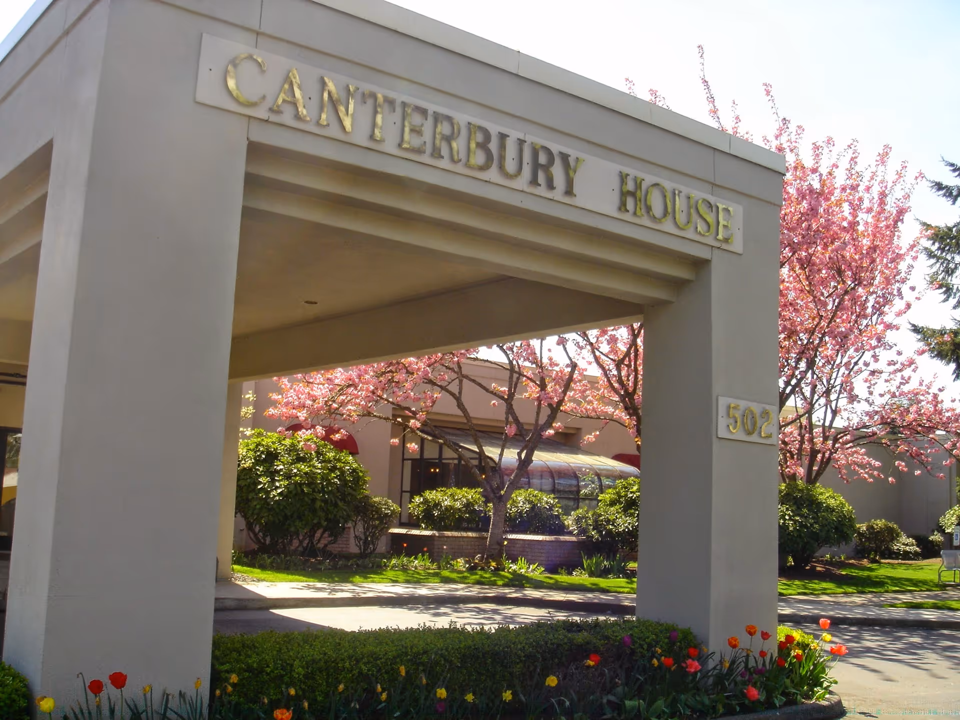 Entrance archway of Canterbury House with the name displayed on top and the number 502 on the side pillar. Surrounding the entrance are blooming pink trees, green bushes, and colorful flowers in a well-maintained garden area.
