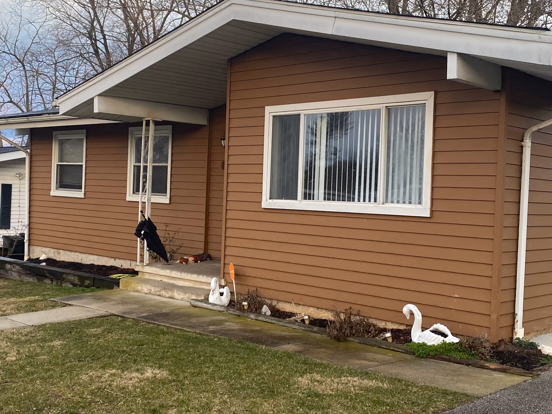 Exterior view of a single-story brown house with white trim. There are three windows visible, a small concrete porch with steps, and two white swan garden ornaments near the foundation. Leafless trees are in the background and a patch of grass is in the foreground.