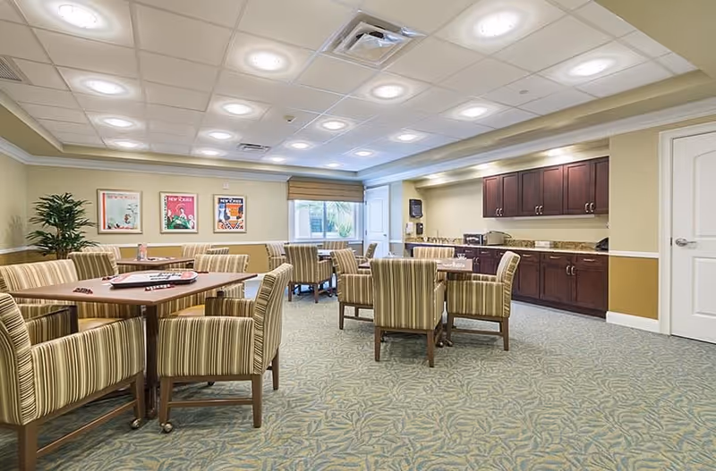 A well-lit common room with multiple tables and striped upholstered chairs arranged for group seating. The room features a carpeted floor with a leafy pattern, beige walls with white trim, and a ceiling with recessed lighting. There is a kitchenette area with dark wood cabinets, a countertop, and appliances along one wall. Three framed colorful posters hang on one wall, and a window with a beige blind allows natural light into the room. A potted plant is placed in the corner.