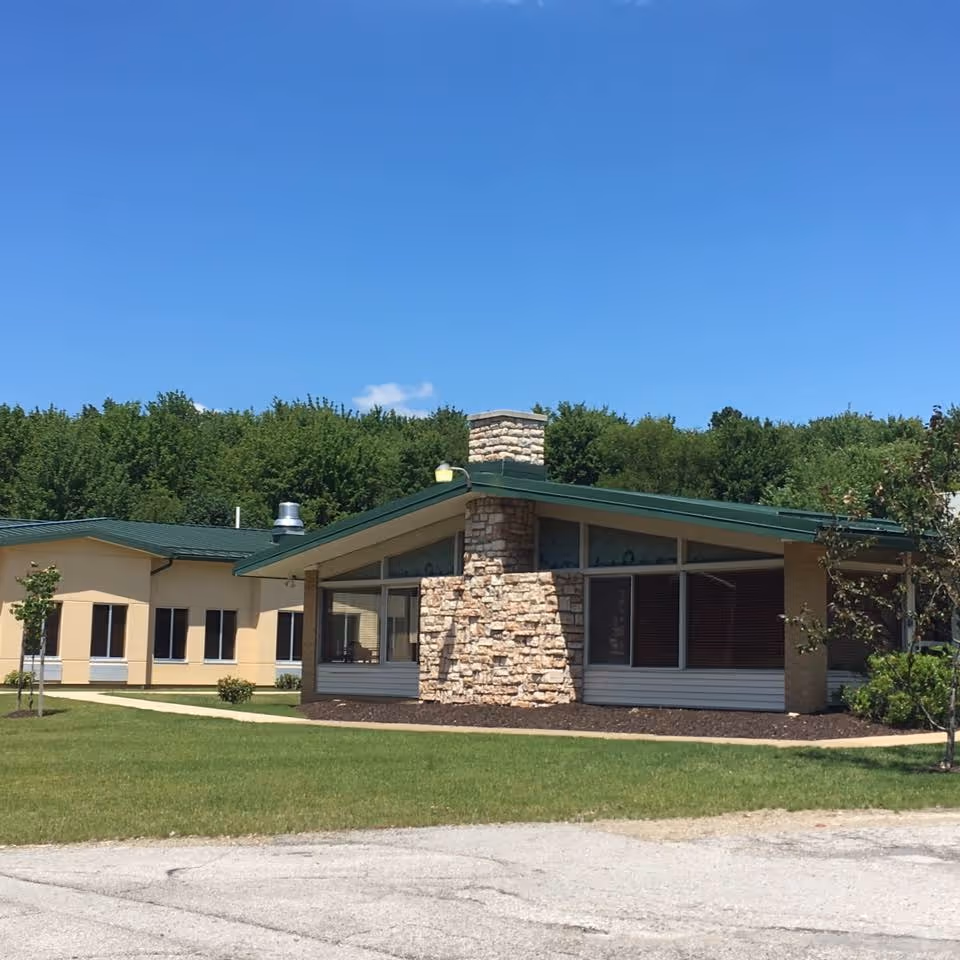 Single-story building with a green roof, stone chimney and large windows set on a grassy lawn under a clear blue sky.