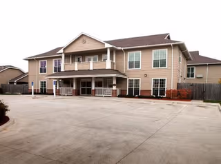 Two-story beige assisted living building with a covered entrance, balcony, and an empty concrete parking lot in front.