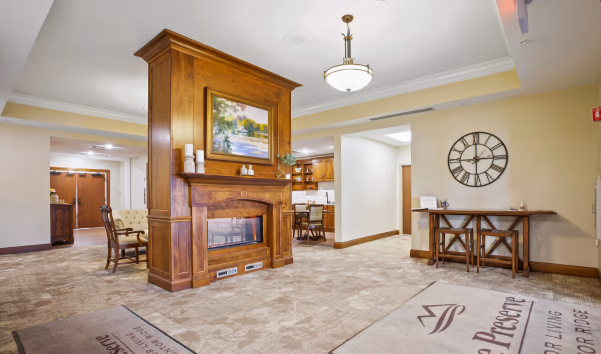 Warm, open senior living lobby with a central wooden fireplace, seating areas, and a wall clock.