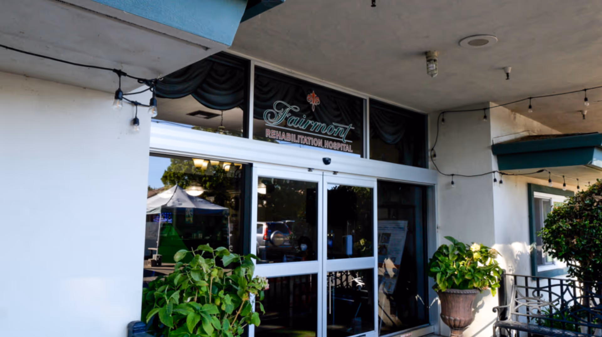 Entrance to Fairmont Rehabilitation Hospital with glass sliding doors, potted plants on either side, string lights hanging from the ceiling, and a sign above the door displaying the facility name.