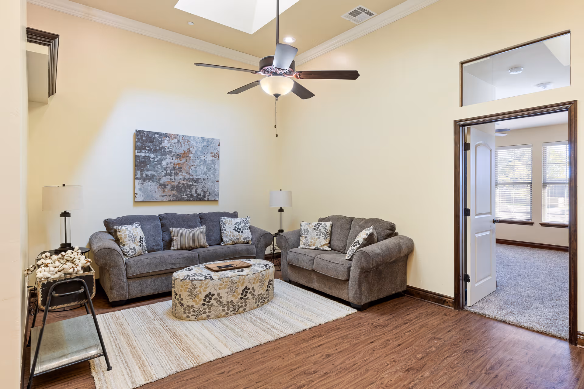 A cozy living room with two gray sofas adorned with patterned pillows, a matching oval ottoman, a light-colored area rug, two floor lamps, a piece of abstract wall art, and a ceiling fan with a light. The room has light yellow walls, wood flooring, and an open doorway leading to a carpeted room with large windows.