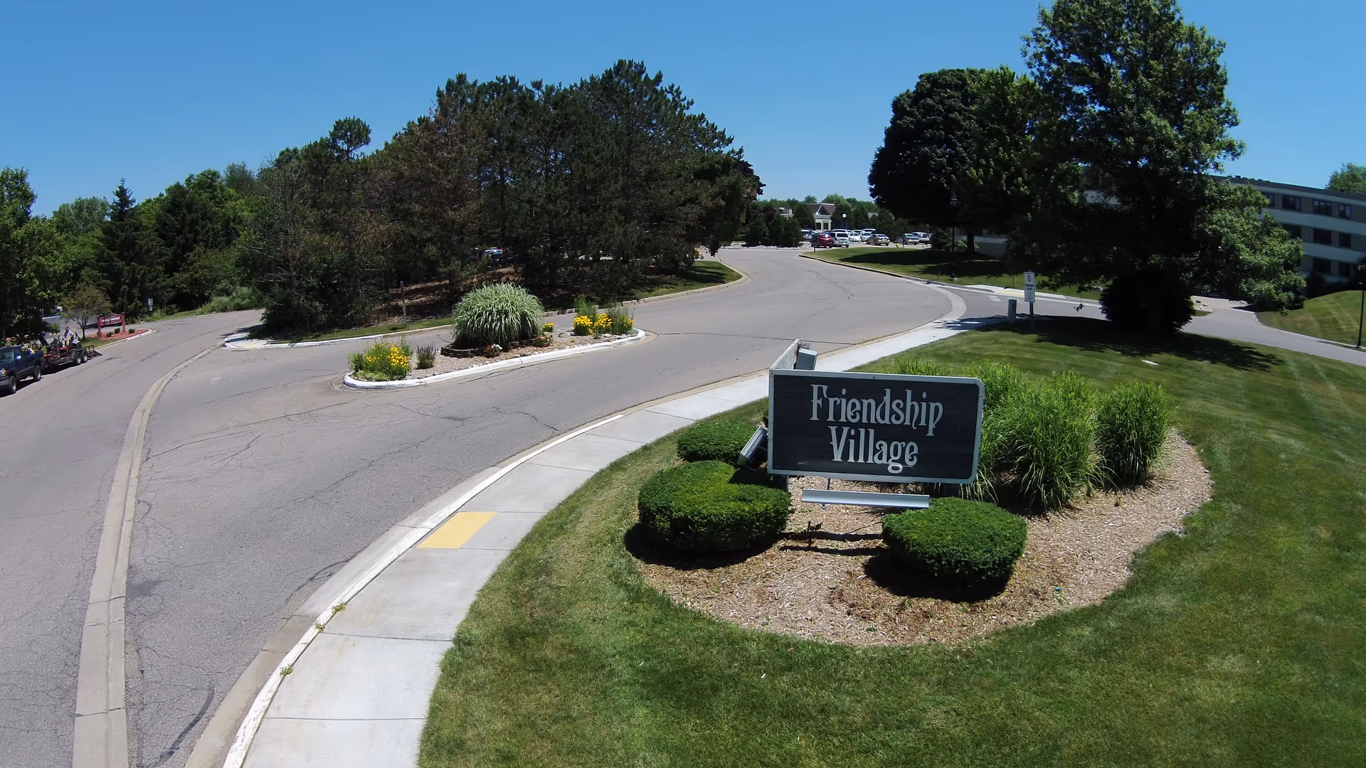 Outdoor view of the entrance area to Friendship Village Kalamazoo, showing a landscaped roundabout with a sign reading 'Friendship Village' surrounded by bushes and grass, with trees and a building in the background under a clear blue sky.