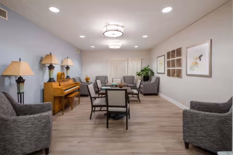 Communal living room with chairs and small tables, a piano, lamps, and framed wall art.