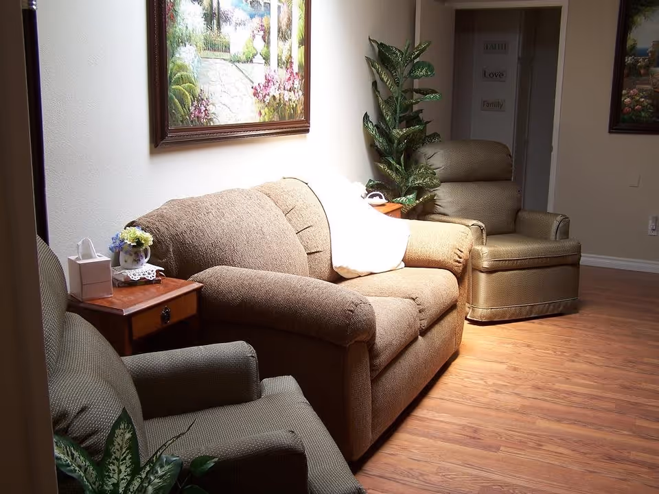 A cozy living room area with a brown upholstered sofa, two armchairs, a wooden side table with a tissue box and a small flower vase, a potted plant, and framed paintings on the walls. The floor is wooden, and there is a hallway visible in the background with wall decor that reads 'Faith, Love, Family'.