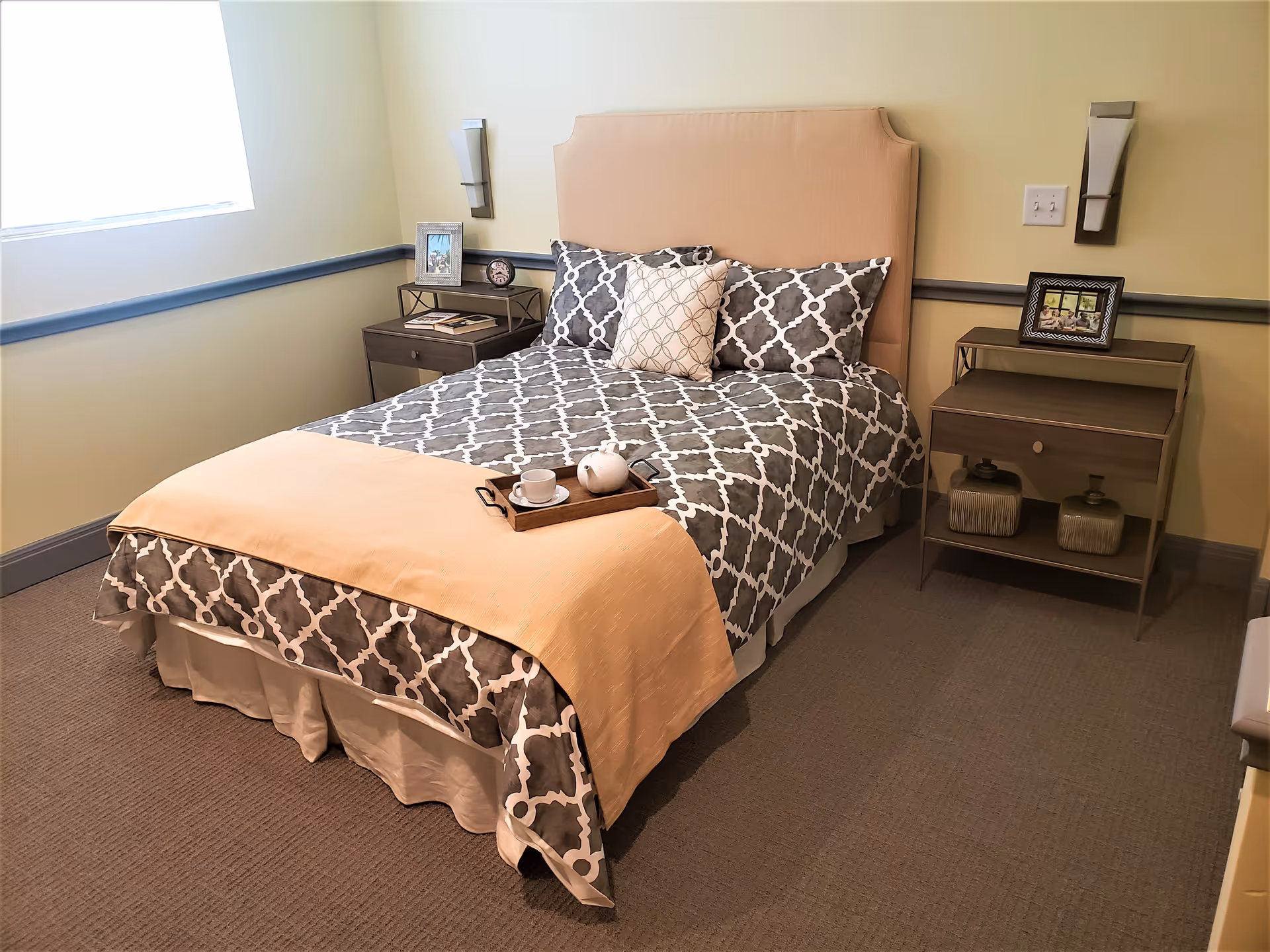A neatly made bed with a beige headboard and patterned gray and white bedding, accented with a light peach throw blanket. There are two matching nightstands on either side of the bed, each with decorative items including framed photos and small vases. The room has beige walls with a gray chair rail and carpeted floor, and a window on the left side letting in natural light.