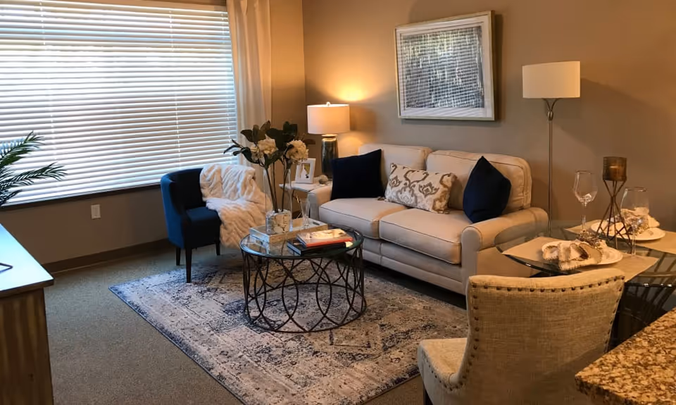 A cozy living room with a beige sofa adorned with decorative pillows, a blue armchair with a white throw blanket, a round glass coffee table with books and flowers, a floor lamp, a table lamp on a side table, and a glass dining table set with plates and wine glasses. The room has a large window with blinds and a framed artwork on the wall.