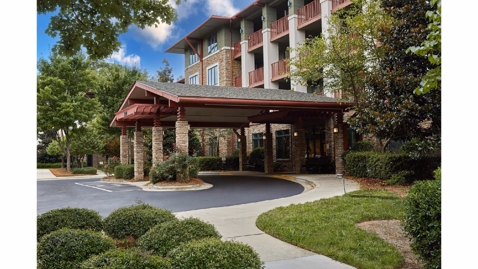 Entrance of a senior living facility named Magnolia Glen featuring a covered drop-off area with stone pillars and a curved driveway. The building has multiple floors with balconies and is surrounded by well-maintained landscaping including bushes, trees, and grass under a partly cloudy sky.