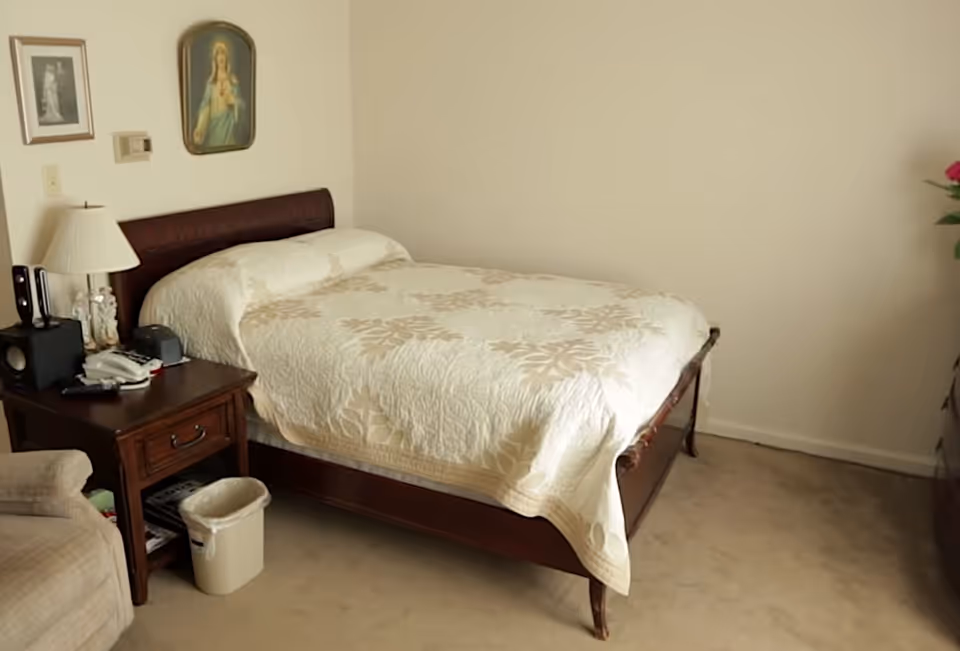 A small bedroom with a single bed covered with a white and beige floral quilt. Next to the bed is a wooden nightstand with a lamp, telephone, and other small items. There is a beige armchair partially visible on the left side. The walls are plain and light-colored, with two framed pictures hanging above the bed. A small trash bin is placed on the floor beside the nightstand.