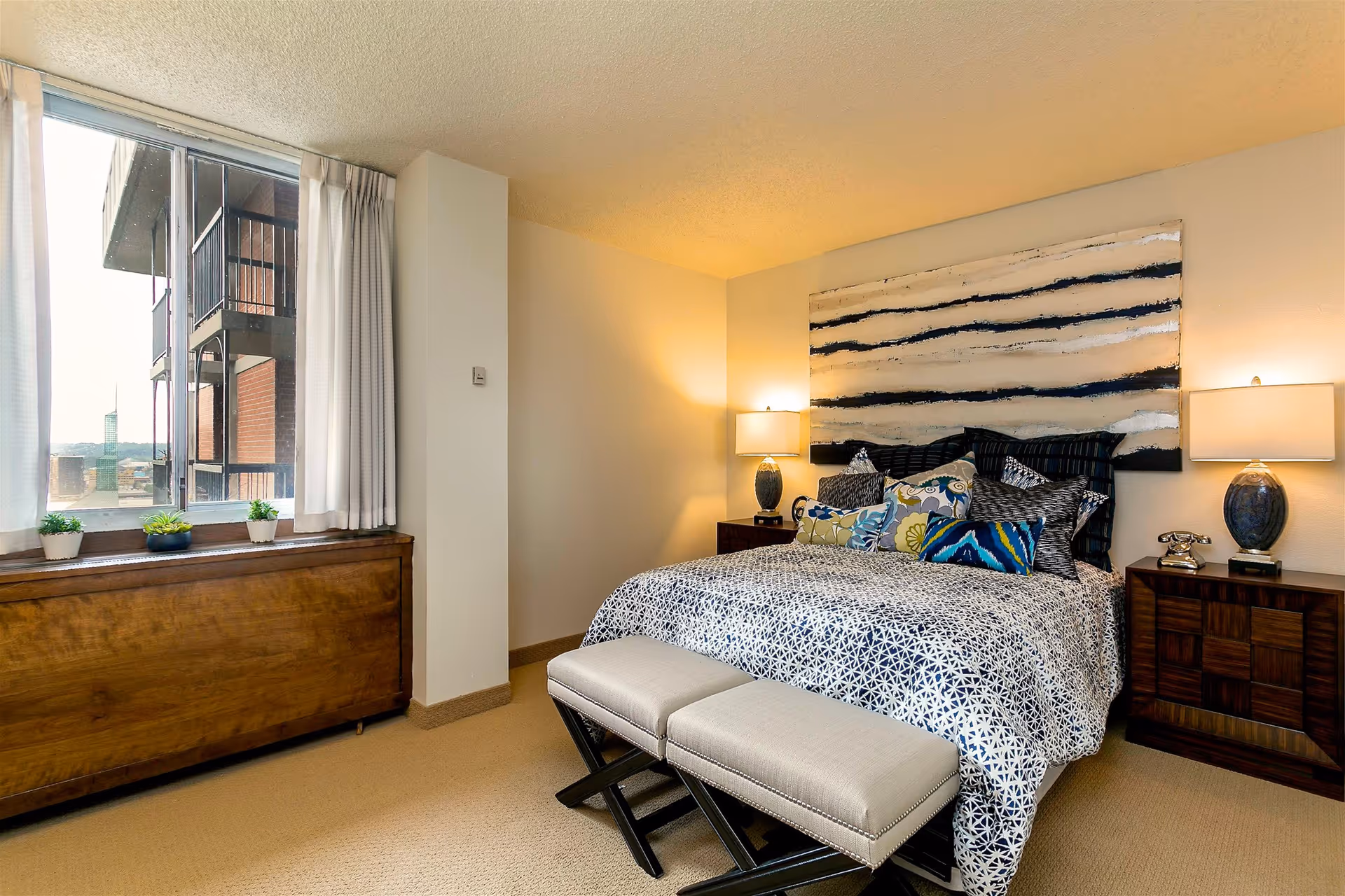 A well-lit bedroom with a large bed adorned with multiple decorative pillows and a patterned bedspread. Two matching bedside tables with blue lamps and a vintage-style telephone are on either side of the bed. Above the bed hangs a large abstract painting with horizontal black and white stripes. A window with white curtains lets in natural light, and a wooden cabinet below the window holds three small potted plants. Two cushioned benches are placed at the foot of the bed.