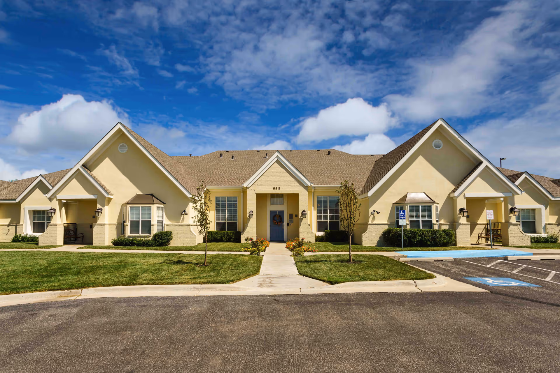 Exterior view of a single-story residential building with a beige facade, multiple peaked roofs, and a central entrance with a blue door. The building is surrounded by a well-maintained lawn, small trees, and a parking area with handicap parking spaces. The sky is partly cloudy with blue patches.