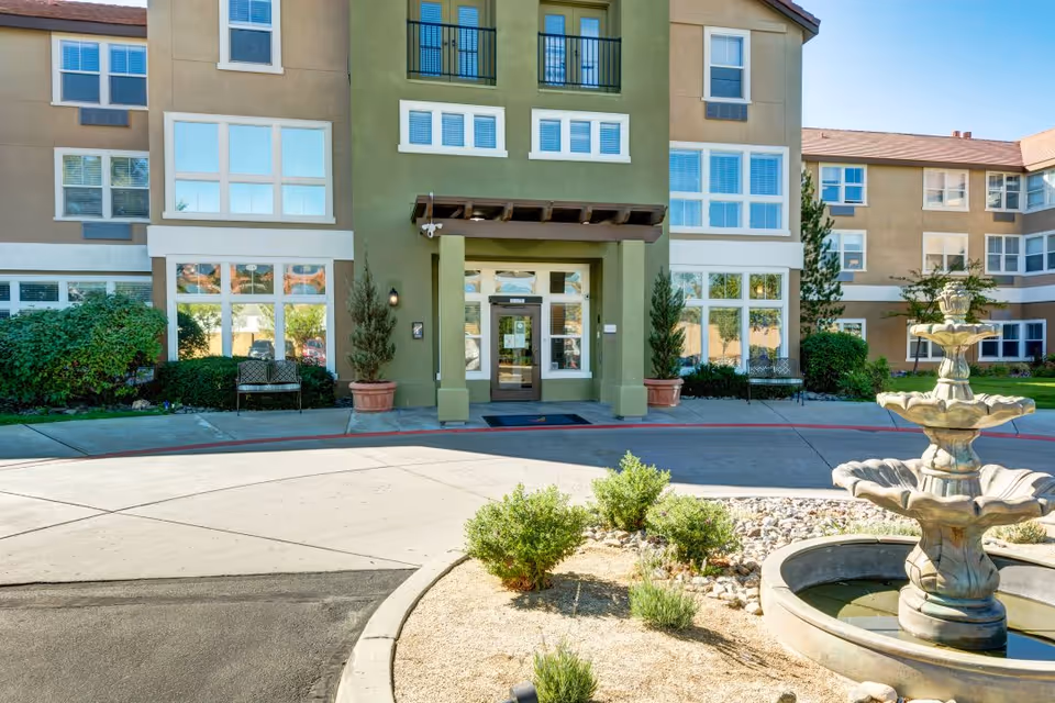 Front exterior view of The Seasons of Reno senior living facility with a three-tiered water fountain in the foreground, surrounded by small bushes and landscaping. The building has multiple windows, a covered entrance with two potted plants, and a bench near the entrance.