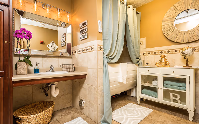 A well-decorated bathroom featuring a sink with a large mirror above it, a purple orchid plant, and a soap dispenser. To the right, there is a bathtub partially hidden by light blue curtains, a white cabinet with glass doors containing folded towels, and decorative items including a globe and a golden frog figurine. The walls are tiled halfway up with a beige and brown diamond pattern, and the floor has matching tiles with two white patterned rugs.