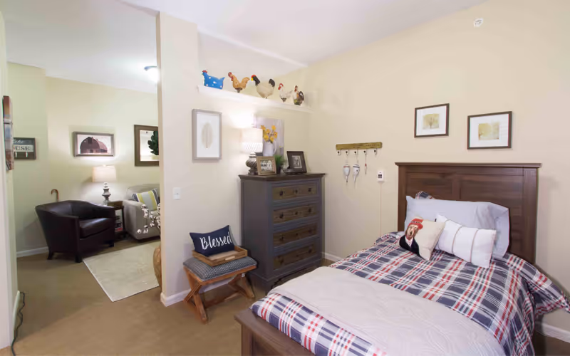 A cozy bedroom in a senior living facility with a wooden bed featuring plaid bedding and decorative pillows. Next to the bed is a dark wooden dresser with framed pictures and a lamp on top. Above the dresser is a shelf with decorative ceramic chickens. A small bench with a cushion and a pillow that says 'Blessed' is placed beside the dresser. The room opens into a living area with a leather armchair, a side table with a lamp, and framed artwork on the walls.