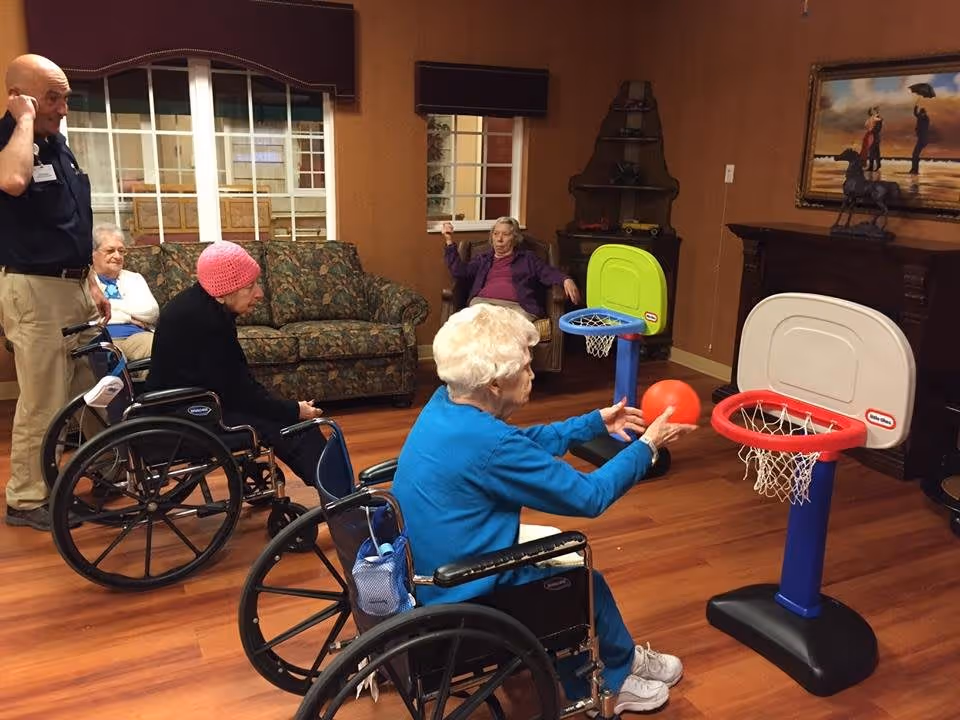 A group of elderly individuals in a common room at Cobblestone Crossings. Two women in wheelchairs are playing with small basketball hoops, one holding a red ball aiming to shoot. Another elderly woman sits on a couch, and a man stands nearby observing. The room has wooden floors, a floral couch, a painting on the wall, and warm-colored walls.