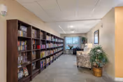 Interior view of a senior living facility hallway featuring a long bookshelf filled with books on the left side, a seating area with a patterned armchair and a lamp on the right, a potted plant in a wicker basket, and a window at the far end letting in natural light.