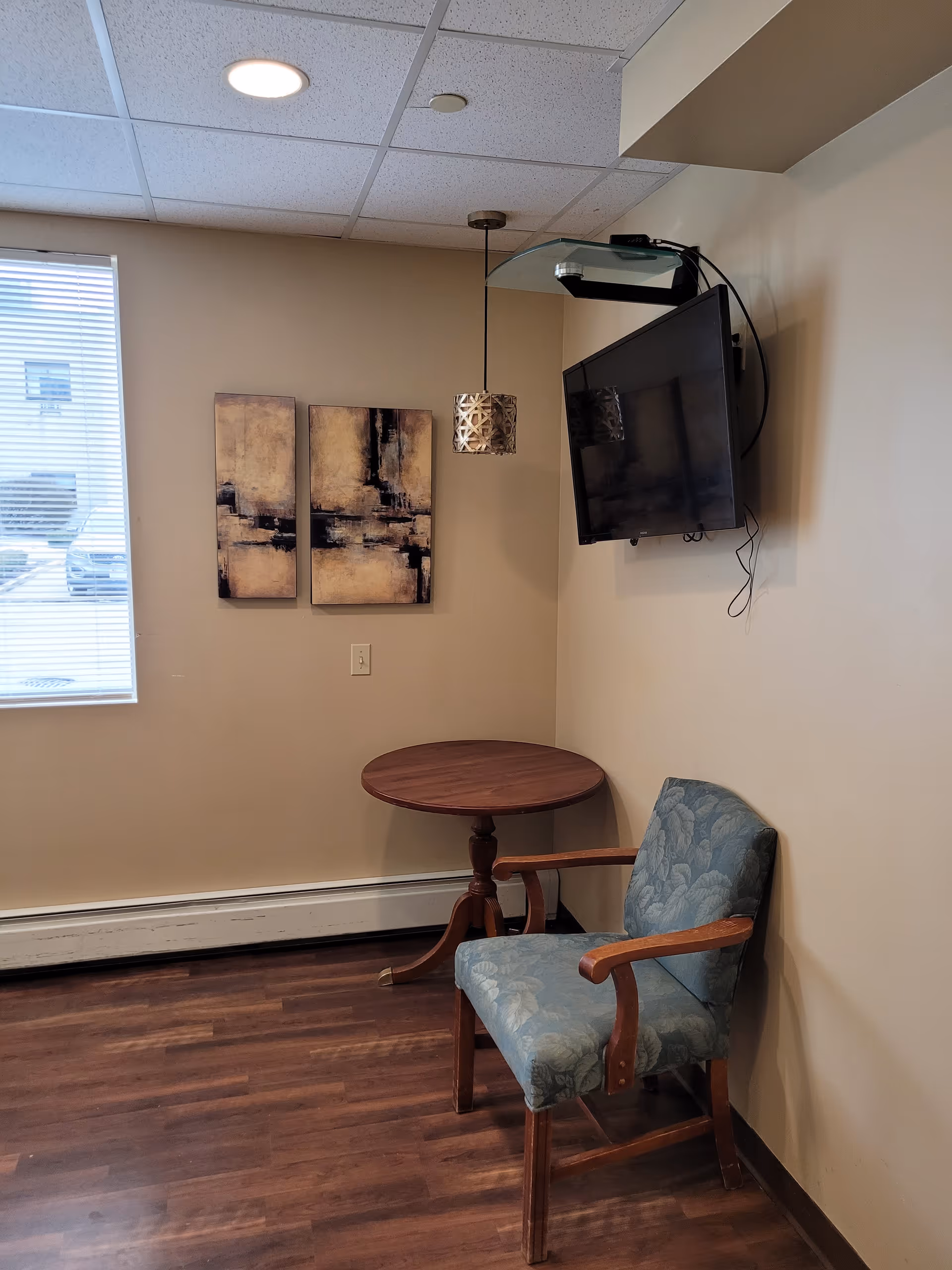 A small corner of a room with a wooden round table, a blue upholstered armchair, a wall-mounted flat screen TV, two abstract paintings on the wall, and a window with blinds letting in natural light.