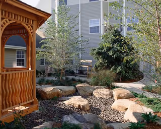 A landscaped courtyard with a wooden gazebo, large decorative boulders, trees, and the exterior of an apartment building in the background.