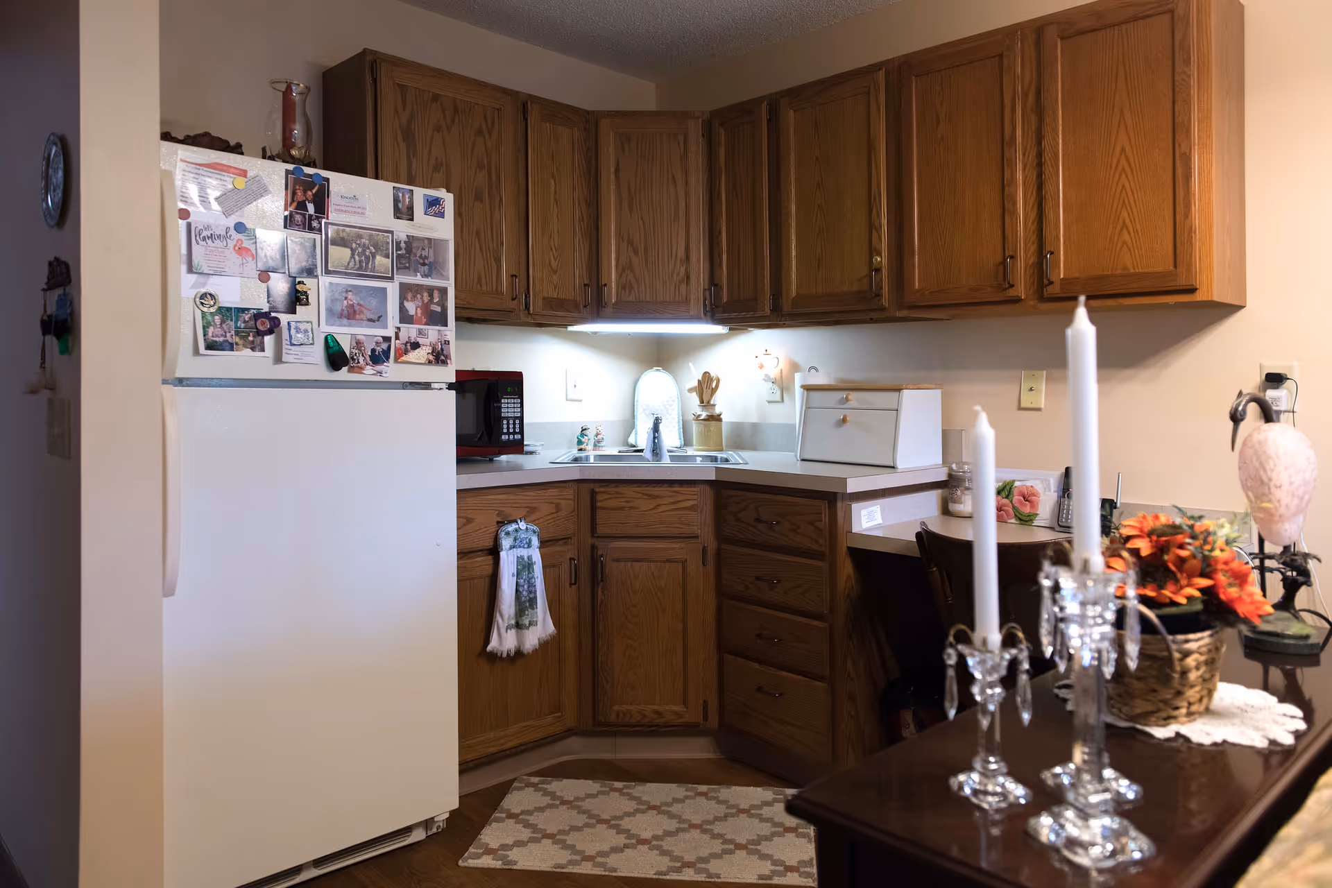 A cozy kitchen corner with wooden cabinets, a white refrigerator covered with photos and magnets, a microwave on the counter, a double sink with a small lamp above it, and a patterned rug on the floor. In the foreground, there is a table with two white candles in crystal holders and a basket of orange flowers.