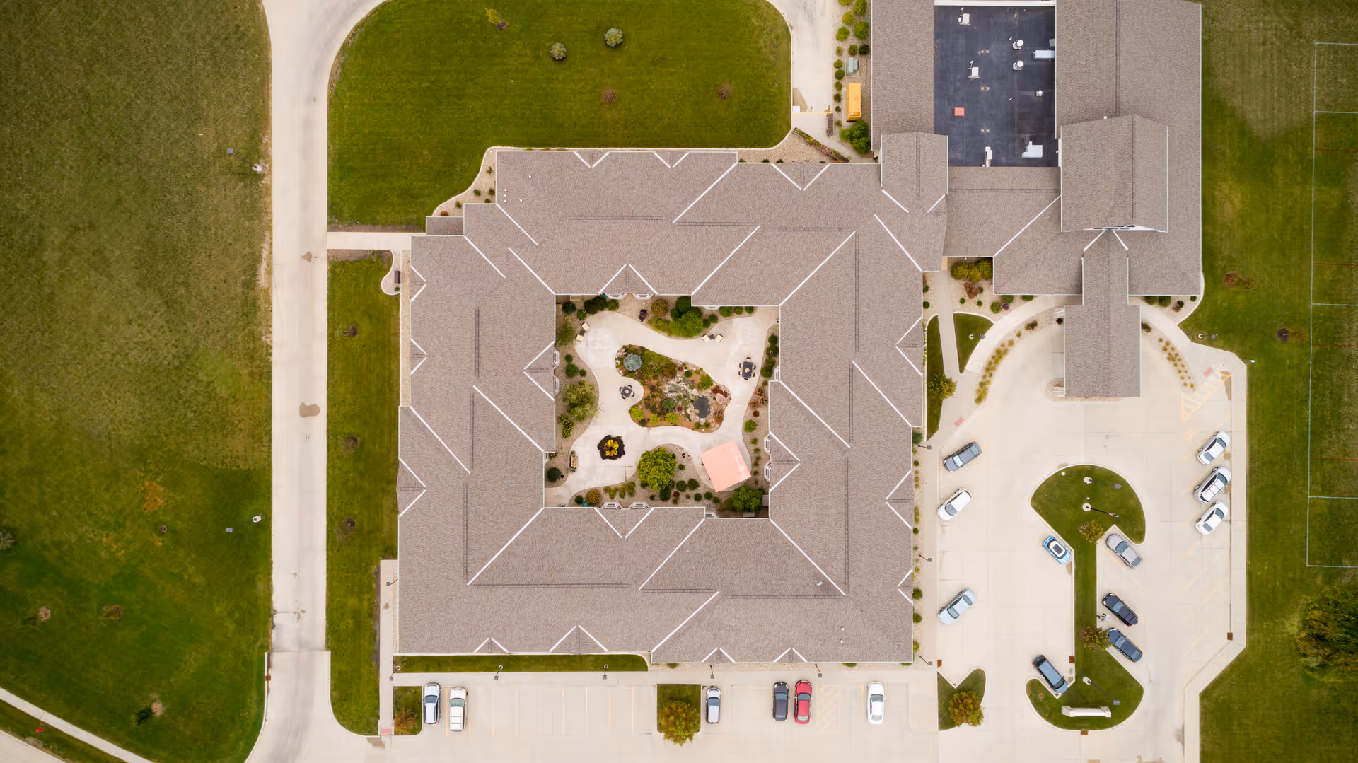 Aerial view of The Village at Mercy Creek senior living facility showing a large square building with a central courtyard garden. Surrounding the building are parking lots with several cars and green grassy areas.