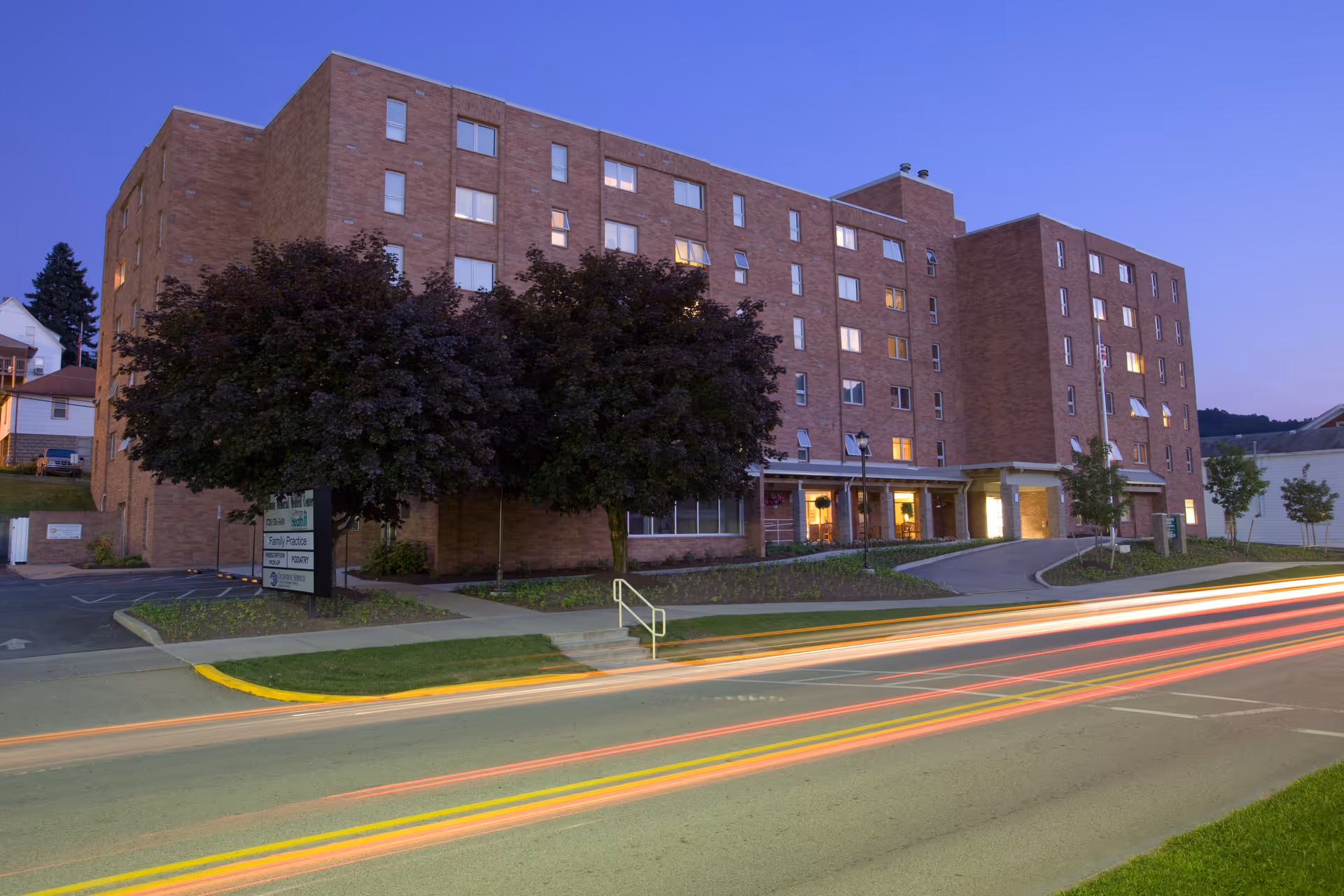 Exterior view of a multi-story brick building at dusk with lit windows and a driveway entrance. Trees and a sidewalk are visible in front of the building, with light trails from passing vehicles on the road in the foreground.
