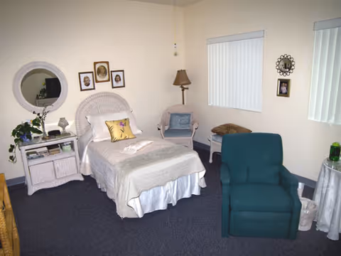 A cozy bedroom in an assisted living facility featuring a single bed with white bedding and a decorative pillow, a wicker nightstand with a plant and lamp, a wicker chair with a cushion, a green upholstered armchair, and framed pictures on the walls. The room has carpeted flooring and two windows with vertical blinds.