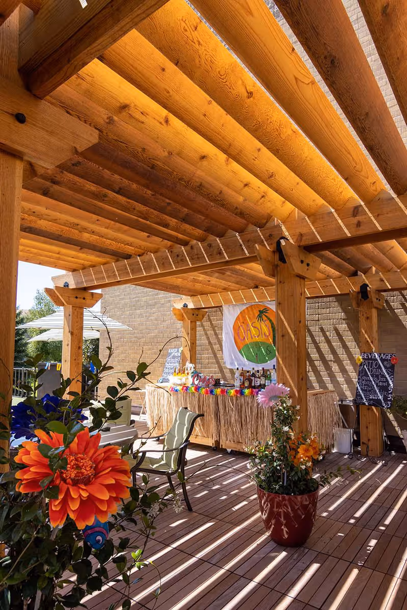 Outdoor patio area with a wooden pergola overhead casting shadows on the wooden deck floor. There are colorful flowers in pots and a tiki-style bar decorated with a grass skirt and a banner that reads 'OASIS'. A chalkboard sign lists beverage and patio drink options. Patio chairs and umbrellas are visible in the background.