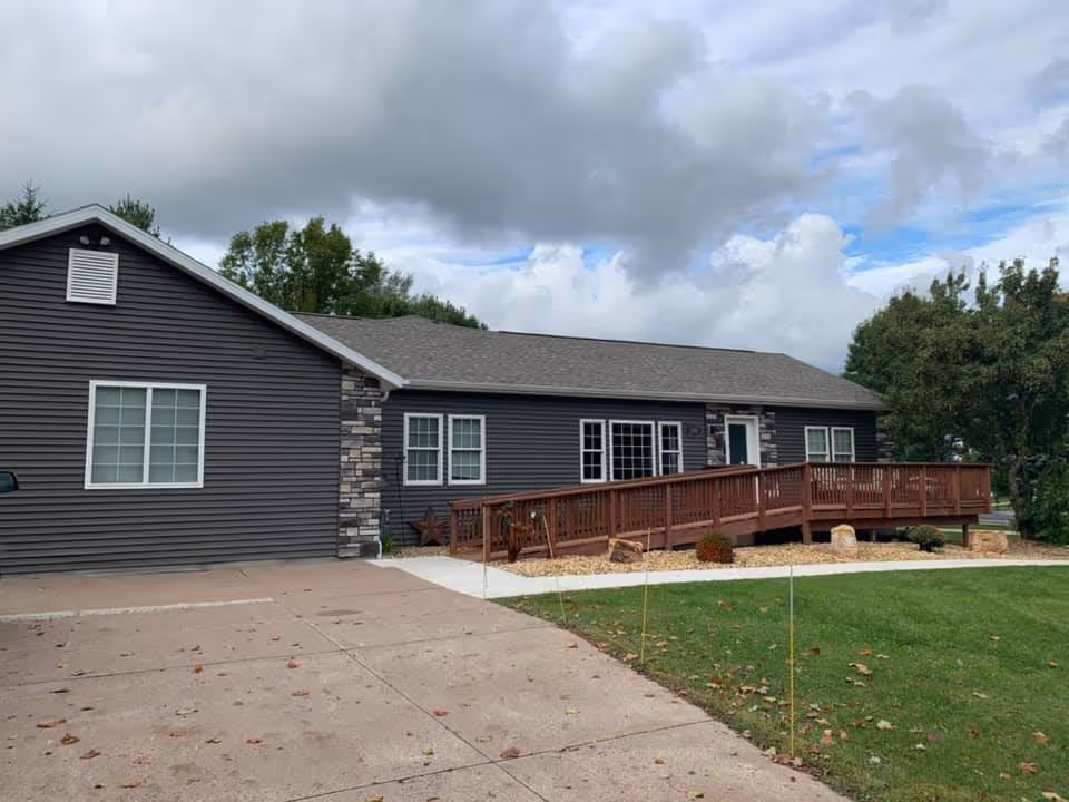 Exterior view of a single-story building with dark gray siding and stone accents. The building has multiple windows and a wooden ramp leading to the entrance. There is a concrete driveway and a well-maintained lawn with some trees and landscaping. The sky is partly cloudy.